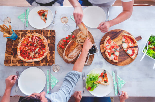 Famille ou amis assis à une table à manger avec plats à emporter. Il y a de la pizza et de la salade sur la table avec du pain. Il y a quatre couverts. Forme de prise de vue en grand angle ci-dessus